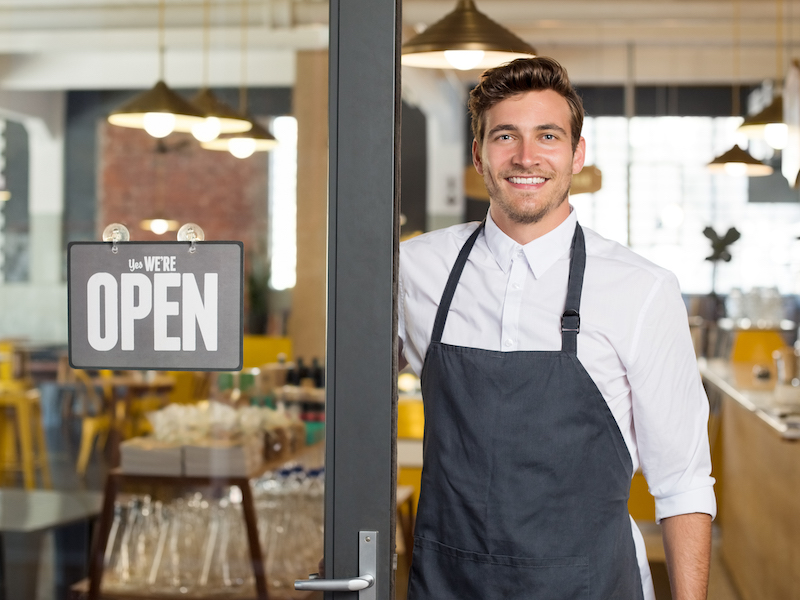 Man in an apron standing in a doorway to a business with a 'We're open' sign on the window.